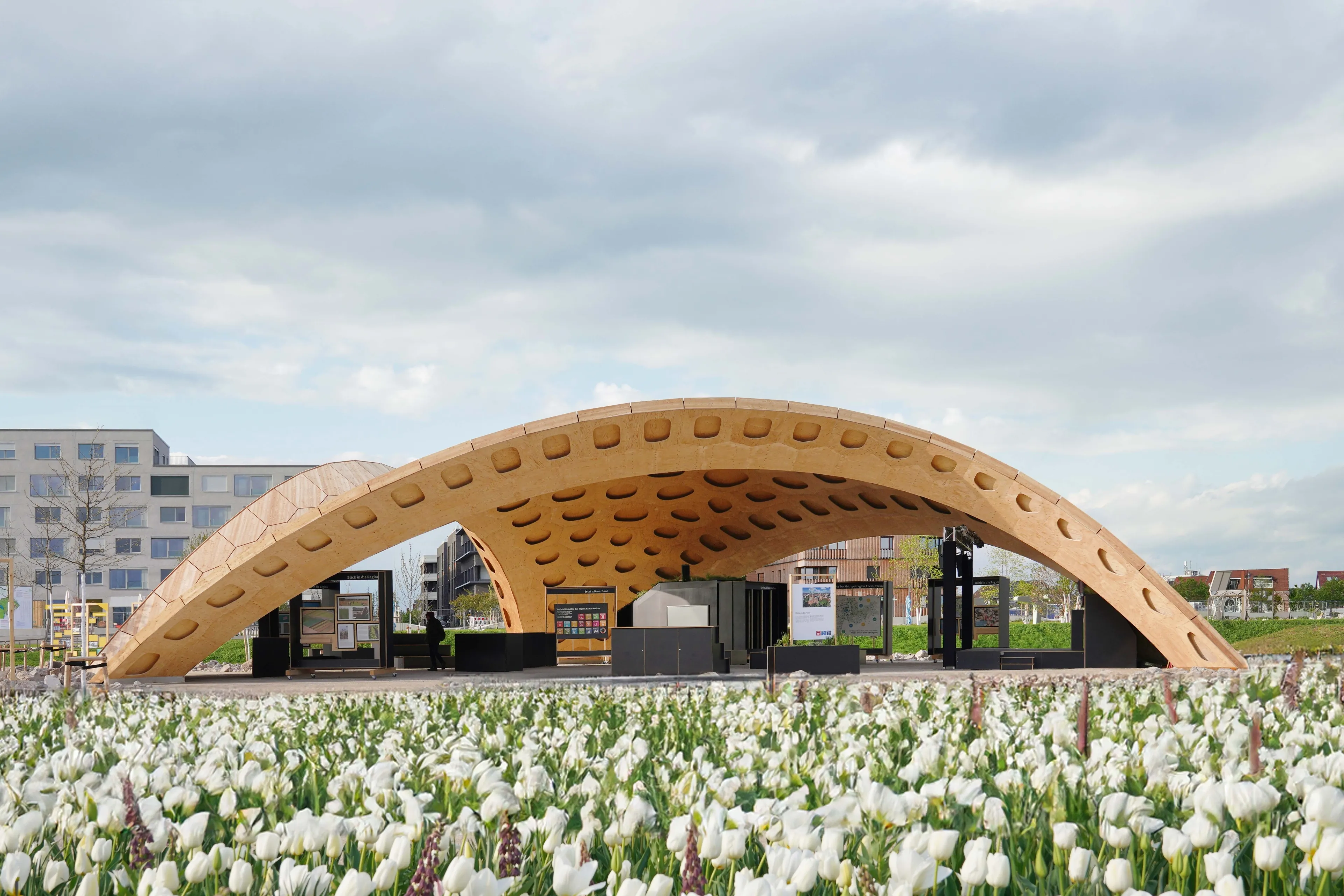 BUGA Wood Pavilion remontado em Mannheim (2023), arco de painéis hexagonais de madeira em paisagem urbana