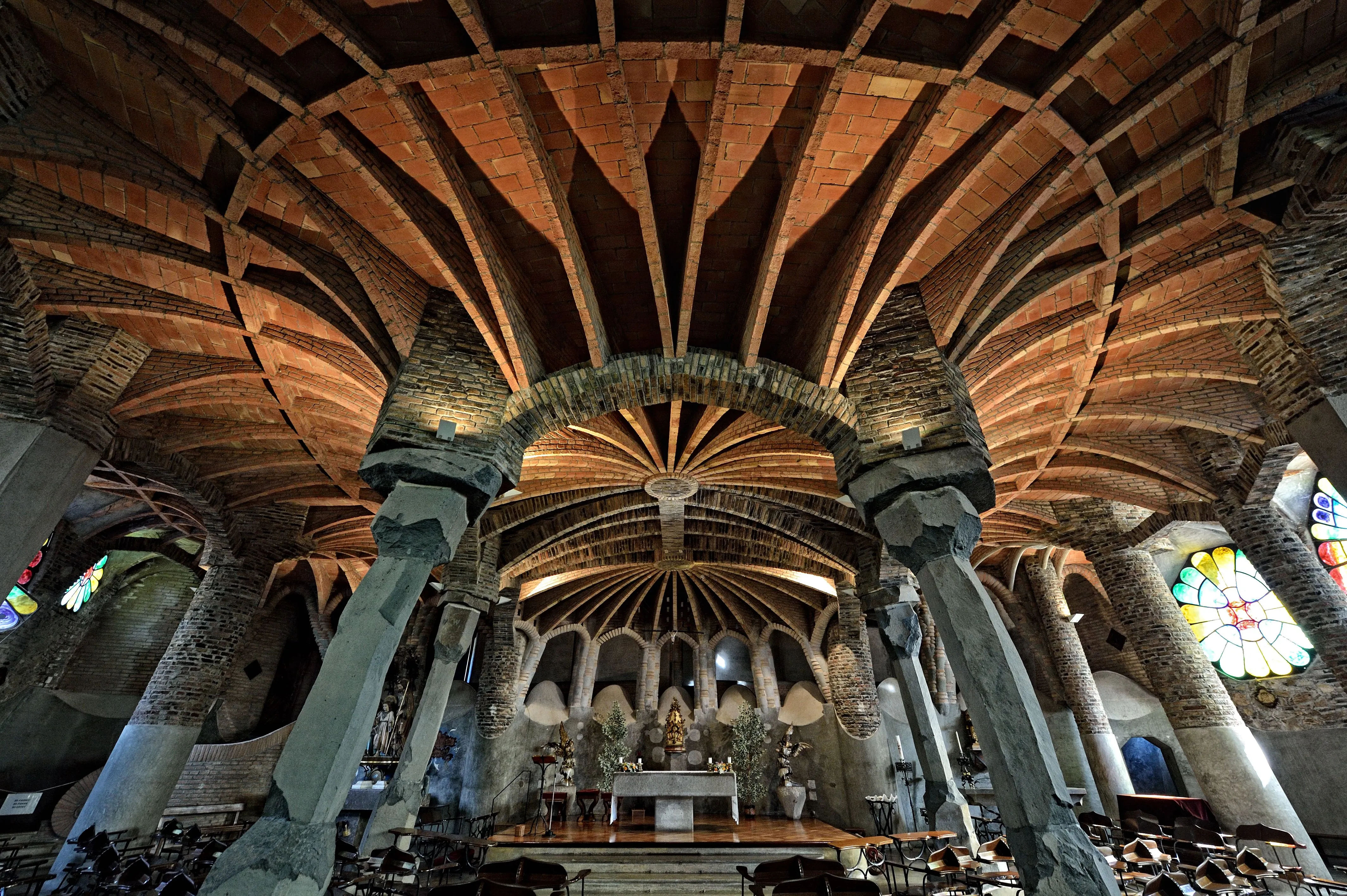 Vista axial do interior da Cripta da Colônia Güell, mostrando a abóbada de tijolo nervurada em leque, colunas inclinadas de basalto e altar ao fundo