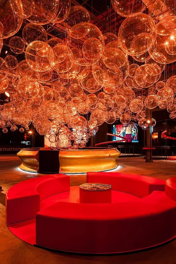 Foto einer Lounge im Vordergrund mit roten, runden Polstersitzen und der goldenen Bar im Hintergrund.