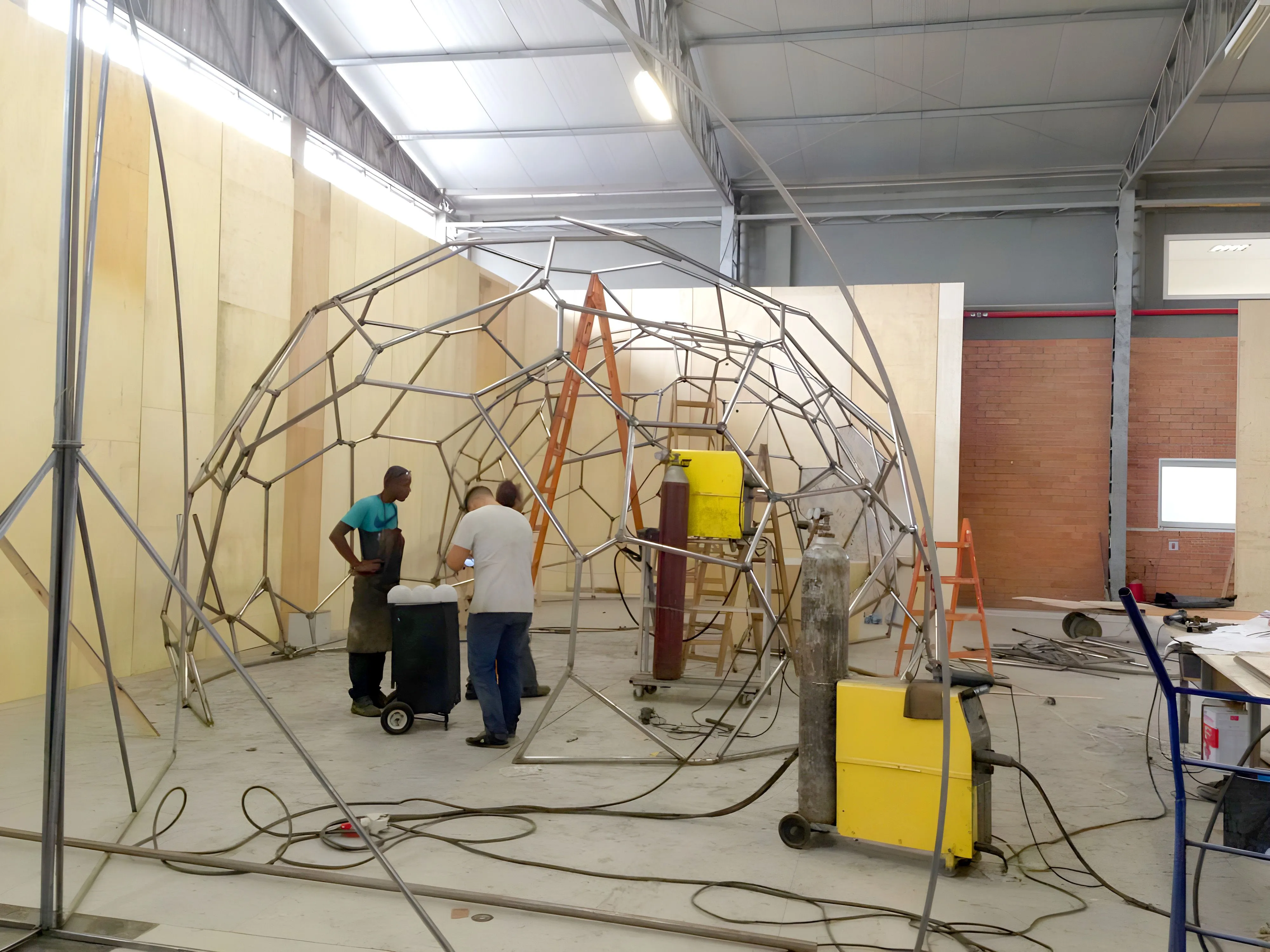 Fabrication of pavilhão O3 by Paleta Stands. A blacksmith takes photos of the small scale model, while two other appear in the background.