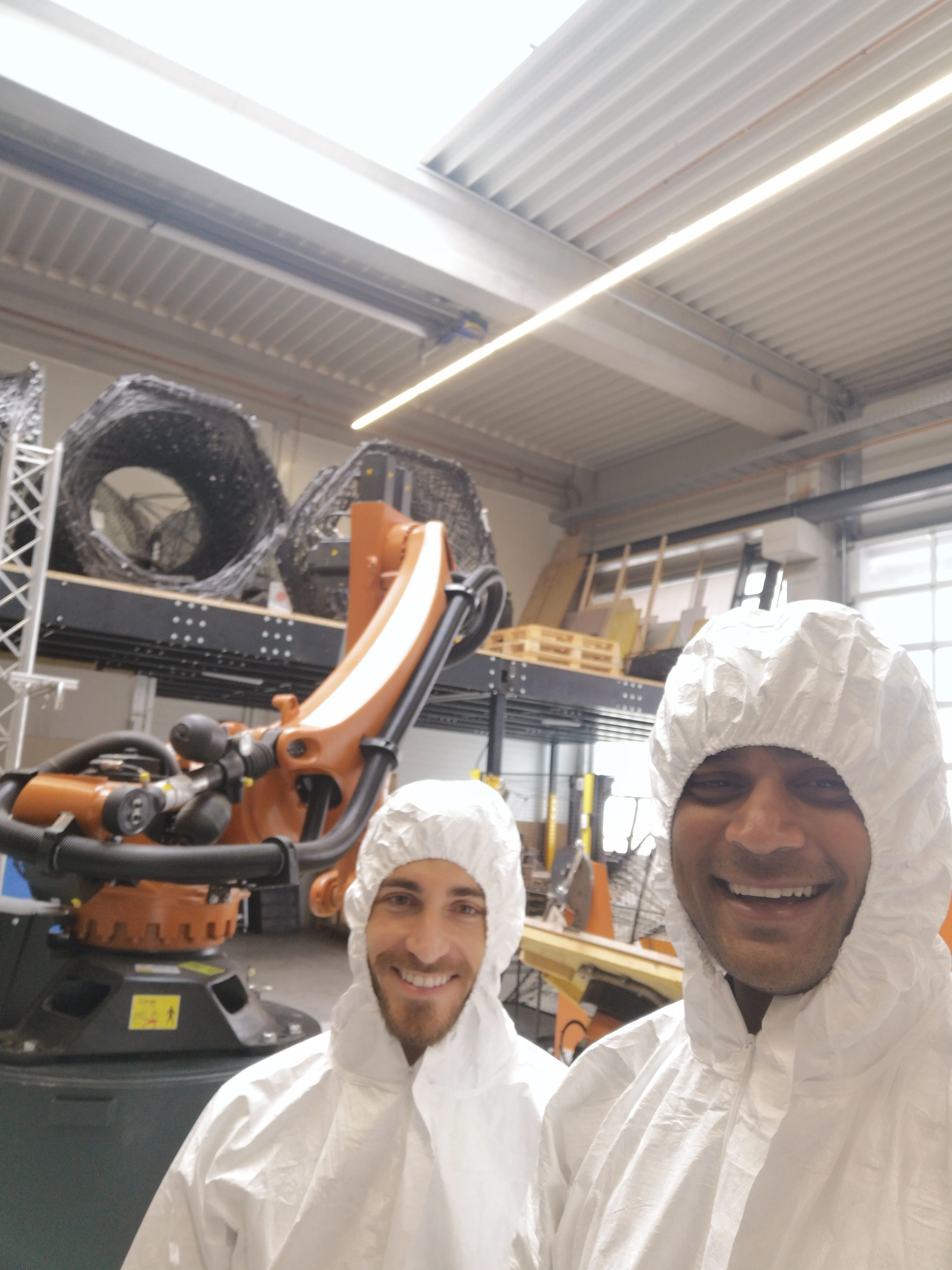 Daniel Nunes Locatelli and Anand Shah in protective suits at the ICD lab, with the KUKA robot and previous filament-wound specimens visible in the background.