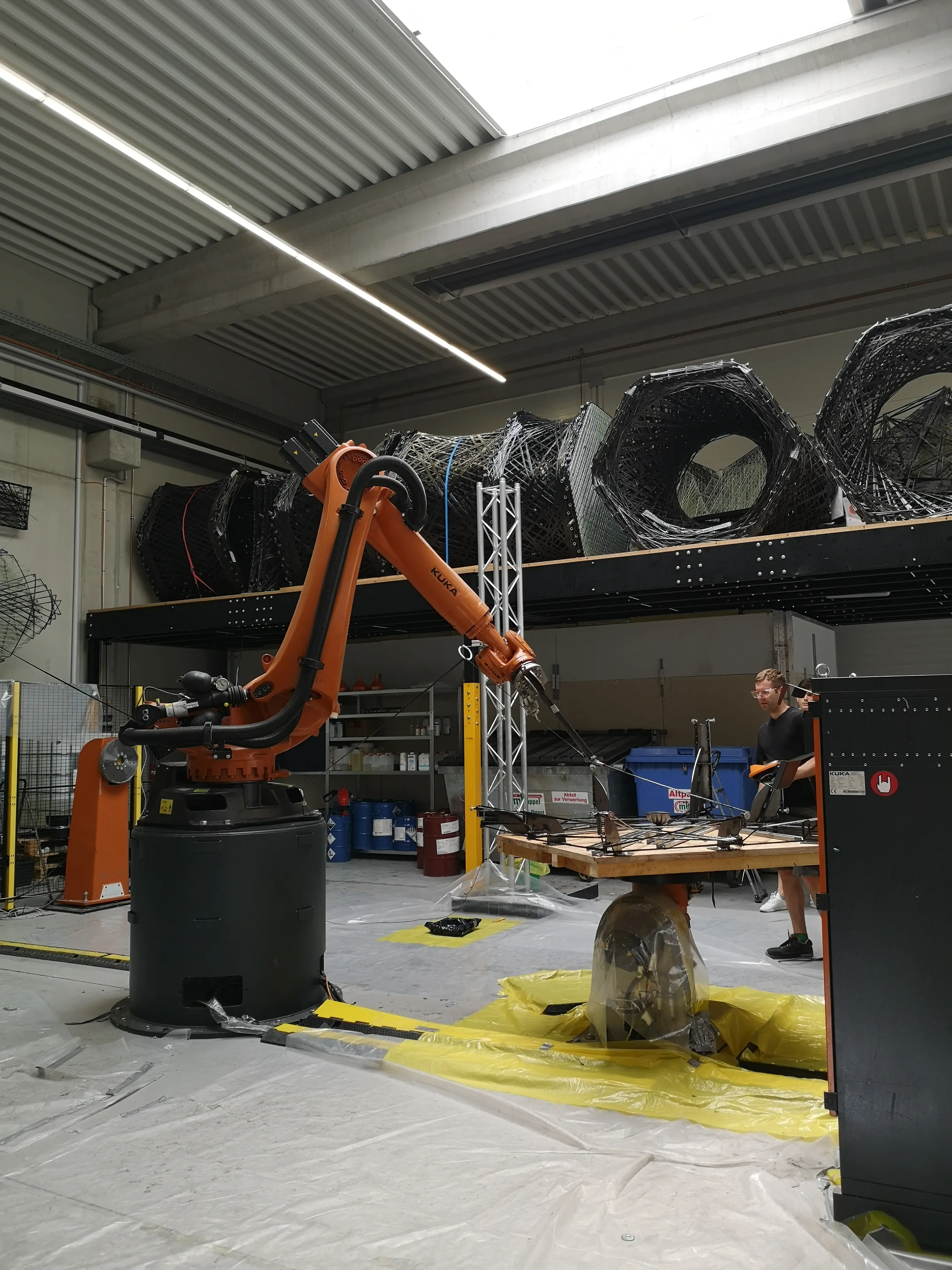The KUKA robotic arm in the lab with carbon fiber winding samples stored on shelves above.